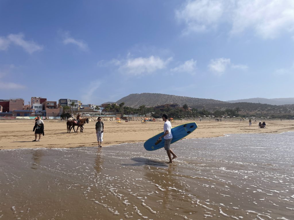 Ein Surfer geht mit seinem Surfbrett am Strand von Taghazout in Marokko entlang; im Hintergrund erheben sich ein Hügel und ein Dorf.