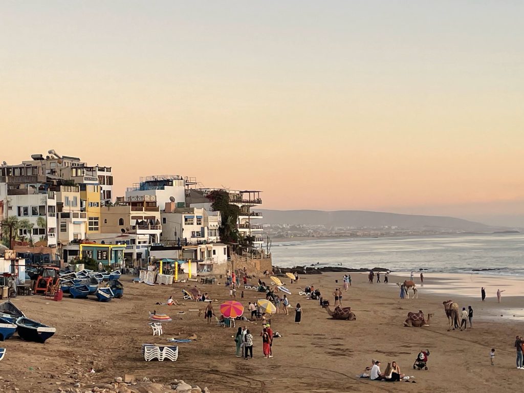 Das Küstendorf Taghazout in Marokko in der Abenddämmerung mit blauen Fischerbooten, bunten Sonnenschirmen und Kamelen am Strand